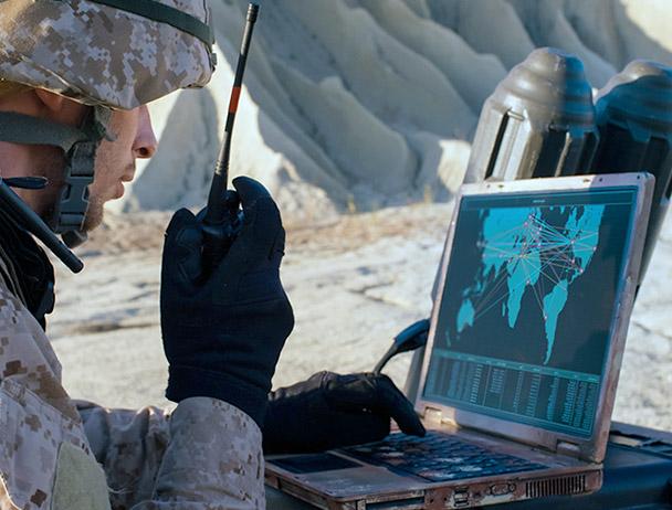 Soldier talking on a handheld satellite radio, looking at a map on a laptop